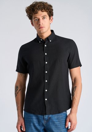 Jeune homme aux cheveux bouclés portant une chemise noire à manches courtes boutonnée et un jean bleu, montrant des tatouages sur les deux avant-bras sur un fond clair.