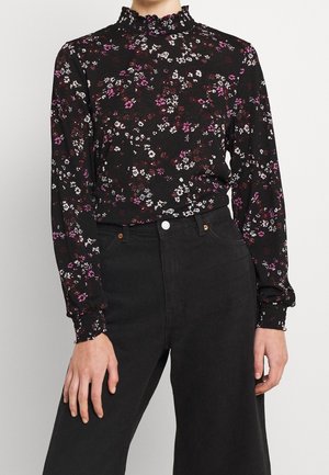 Blusa nera a fiori con motivi rosa e bianchi, presenta un colletto alto con volant e maniche aderenti, abbinata a jeans neri a vita alta.