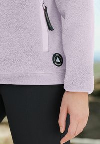 Chaqueta de forro polar de color lila claro con un acabado texturizado, que presenta un bolsillo con cremallera y un acento de logotipo negro redondo en el dobladillo inferior. Leggings negros debajo.