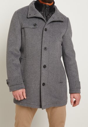 Man draagt een grijze wollen jas met knopen over een zwarte jas met rits en beige broek, staand tegen een eenvoudige lichte achtergrond.