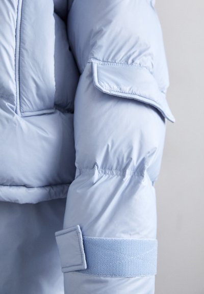 Lichtblauwe pufferjack met een gequiltde textuur, voorzien van een klittenbandsluiting en een patchzakje op de mouw. Zacht, glad materiaal.