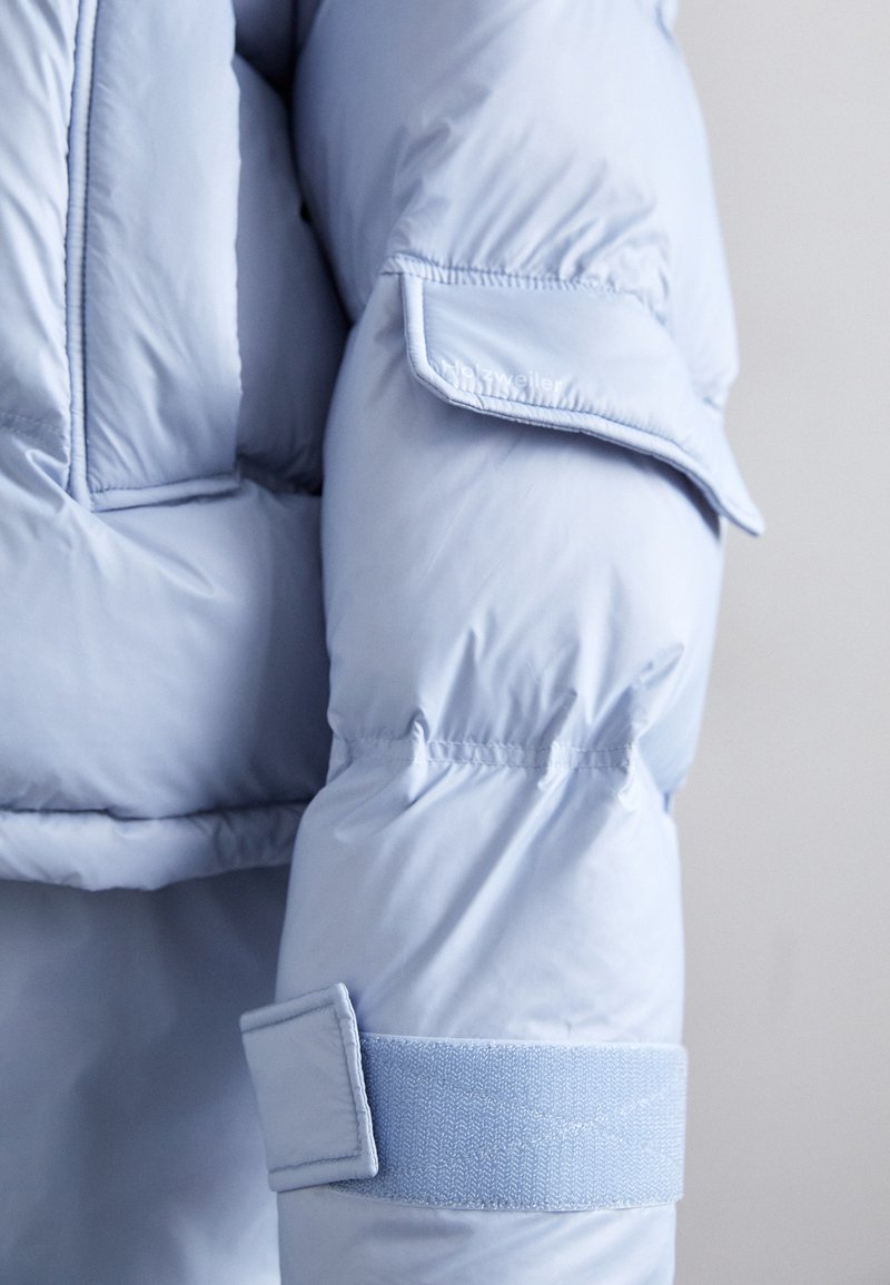 Veste rembourrée bleu clair avec une texture matelassée, dotée d'une sangle Velcro et d'une poche à rabat sur la manche. Matériel doux et lisse.