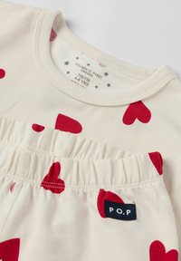 Lichtcrème short-sleeve top en broek set met rode hartpatronen. Inclusief een elastische taille en een marineblauwe P.O.P label.