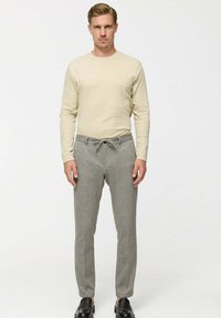 Beige overhemd met lange mouwen en een ronde hals, gecombineerd met grijze tapered broek met een drawstring tailleband. Model draagt donkere schoenen.
