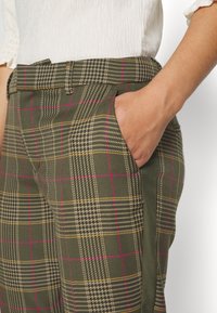 Personne portant un pantalon à carreaux verts avec des lignes roses et jaunes, main dans la poche, associée à un chemisier blanc texturé à manches courtes.