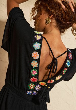 Göndör hajú nő, aki fekete ruhát visel, melyen színes horgolt virágos díszítés és nyitott hát található, fekete zsinórral és bojttal megkötve.