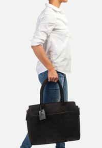 Schwarze Leder-Tote-Bag mit zwei griffen, rechteckiger Form, mit dezenten Nähten und einem kleinen Etikett. Tragend von einer Person in einem weißen Hemd.