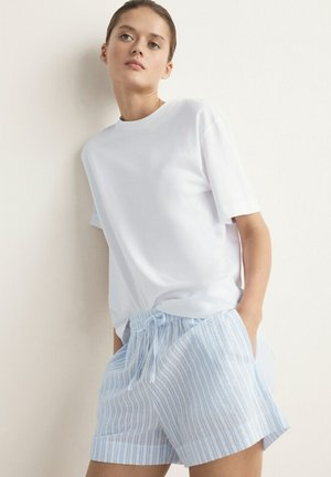 Ung person iført en ensfarvet hvid kortærmet skjorte og lyseblå og hvide stribede shorts med snor i taljen, læner sig op ad en hvid væg.