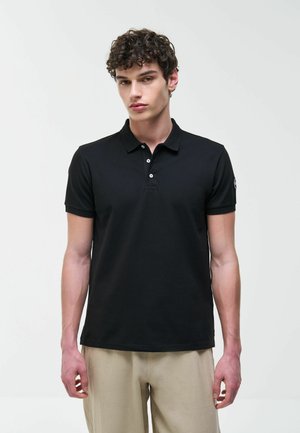 Mladý muž s kudrnatými vlasy, oblečený v černém polo tričku a béžových kalhotách, stojící před jednobarevným bílým pozadím.