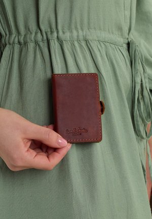 Brown leather wallet with embossed logo, rectangular shape, and visible stitching details, held against a textured green garment.