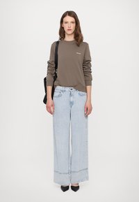 Brun genser med ribbestrikkede mansjetter, lyseblå oversized jeans med frynset kant, og svarte spisse sko. Tote-veske slengt over skulderen.