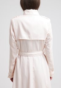 Jasnoróżowy trencz z tylną klapą, długimi rękawami i wiązaniem w talii. Wykonany z gładkiej tkaniny o prostym wzornictwie, bez widocznych wzorów.