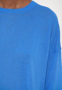 Gros plan d'une personne portant un pull bleu uni à col rond montrant la partie supérieure de la poitrine et la zone des épaules.