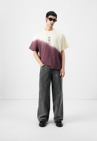 Tricou din bumbac cu un gradient de la crem la burgund, având un imprimeu grafic în centru. Asortat cu blugi gri cu picioare largi și pantofi negri.