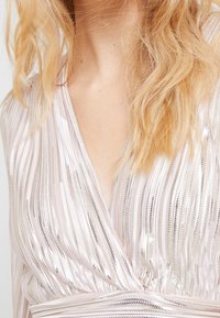 Haut enveloppant scintillant en rose clair avec de fines rayures verticales et des accents métalliques. Dispose d'un décolleté en V profond et d'une taille nouée. Texture douce.