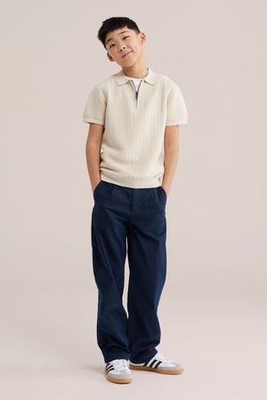 Mlajši deček stoji z rokami v žepih, oblečen v krem polo majico s kratkimi rokavi iz pletenine, temno modre hlače in bele superge s črnimi črtami.