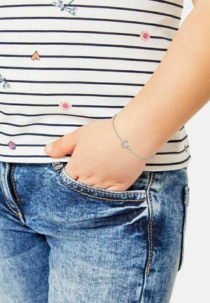 Silberne Kettenarmband mit einem Hufeisenanhänger, am Handgelenk getragen. Die Person trägt ein gestreiftes Shirt in Weiß und Marineblau sowie eine blaue Jeans.