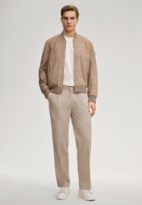 Veste bomber en daim beige avec poignets côtelés, associée à un pantalon beige clair et des baskets blanches. Le mannequin pose sur un fond neutre.