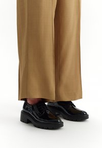 Pantofi din piele patent neagră cu talpă groasă, având șireturi și vârf rotunjit, asortați cu pantaloni largi de culoare bej.