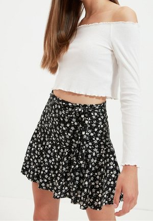 Kvinde iført hvid, skulderfri, langærmet crop top og sort blomsterprintet omslagsnederdel, stående foran en ensfarvet hvid baggrund.