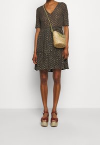 Musta kuviollinen mekko V-pääntiellä ja lyhyillä hihoilla, jossa on kerroksellisia röyhelöitä. Kudottu oljenkudoslaukku vihreällä hihnalla ja punaiset alustakengät.