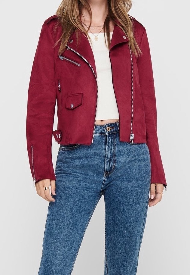Femme portant une veste biker en daim rouge avec des zips argentés, un crop top blanc, et un jean bleu taille haute, debout devant un fond uni.