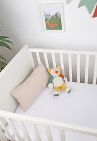 White crib with slatted sides, soft white mattress, beige pillow, and a plush toy, featuring multi-colored accents and textures.