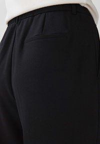 Gros plan sur un pantalon noir sur mesure avec poche passepoilée arrière, porté avec un haut écru.