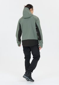 Veste verte résistante à l'eau avec une capuche, des accents noirs et une coupe ample ; portée avec un pantalon noir et des chaussures de sport noires.