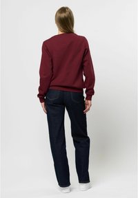 Felpa bordeaux con scollo rotondo, polsini e orlo a costine, abbinata a jeans in denim scuri e sneakers bianche. Tessuto testurizzato, vestibilità casual.