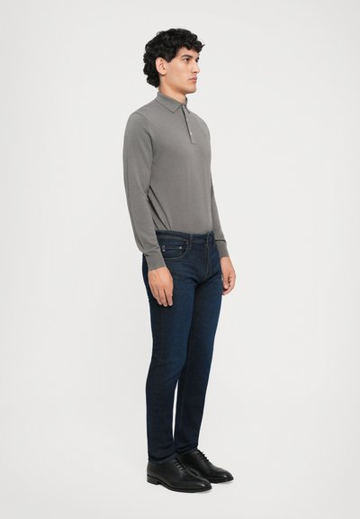 Young man with curly hair standing sideways wearing a grey long-sleeve polo shirt, dark blue jeans, and black dress shoes.