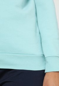 Světle modrá mikina s hladkým povrchem, žebrovanými manžetami a rovnými lemy. Krátký rukáv s volným střihem.