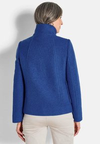 Frau in einem blauen Wollmantel mit hohem Kragen, von hinten gesehen, kombiniert mit hellbeigen Hosen vor einem schlichten weißen Hintergrund.