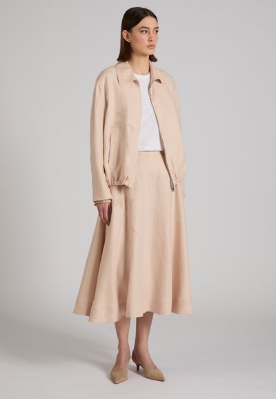 Mujer con chaqueta beige con cremallera, falda midi en línea A a juego, camiseta blanca y tacones beige de punta puntiaguda, posando contra un fondo gris.