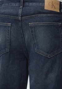 Σκούρο μπλε τζιν με δύο πίσω τσέπες και ταμπά δερμάτινο φιλέτο με ανάγλυφη την ένδειξη "cK" στη μέση.