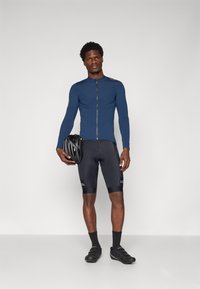 Maillot de cyclisme à manches longues bleu marine avec une fermeture éclair devant, associé à un short rembourré noir. Il tient un casque noir et porte des chaussures de cyclisme noires.