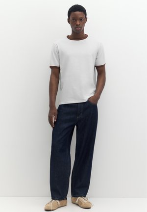 Jeune homme debout contre un mur blanc, portant un t-shirt blanc avec une bordure marron, un jean bleu foncé et des baskets beiges, une main dans la poche.