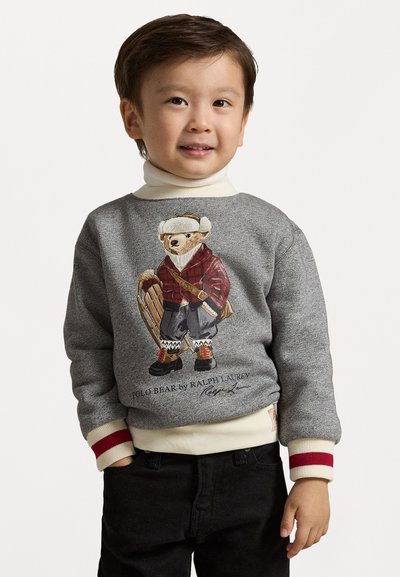 Felpa grigia con un'illustrazione di un orso in abbigliamento invernale, con accenti rossi sui polsini e colletto beige, abbinata a pantaloni neri.