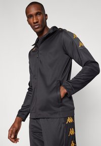 Man stands wearing black zip-up hoodie and matching pants with gold logo stripes on sleeves and pant legs, one hand in pocket.