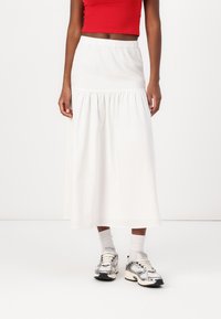 Falda blanca, hasta el tobillo, con una cintura elástica, que presenta un volante fruncido en el dobladillo. Combinada con un top corto rojo y zapatillas grises.