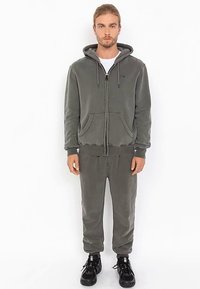 Felpa con cappuccio grigia con zip, coulisse e tasche frontali, abbinata a pantaloni jogger coordinati. Design a lunghezza intera, tessuto morbido e vestibilità comoda.