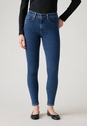 Femme portant un jean bleu ajusté, un haut noir à manches longues et des ballerines noires, debout devant un fond clair uni.