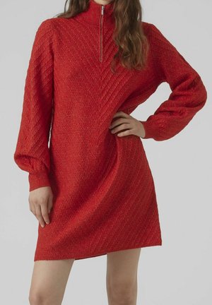 Kvinde iført en rød strikket langærmet kjole med kvartlynlås og chevronmønster, poserer med den ene hånd på hoften.