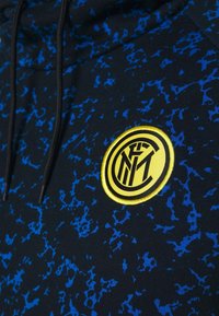 Sweat à capuche noir avec motif éclaboussure bleu, arborant un logo jaune brodé en design circulaire. Cordons de capuche visibles.