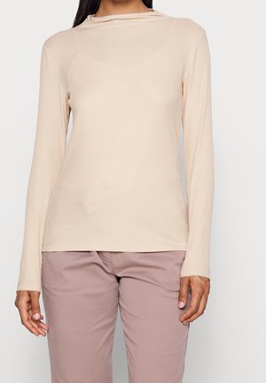 Vrouw die een beige geribd shirt met lange mouwen en mauve broek draagt, staand tegen een effen lichte achtergrond, handen ontspannen langs het lichaam.