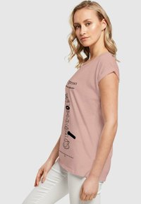 Lyse rosa t-skjorte med korte ermer som har svarte kaffe-relaterte illustrasjoner og tekst på forsiden, laget av mykt bomullsstoff.
