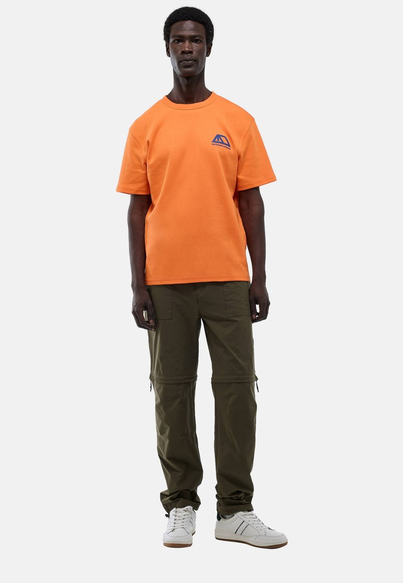 Un homme portant un T-shirt orange avec un logo bleu, un pantalon cargo vert olive et des baskets blanches, se tenant devant un fond uni.
