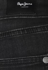 Vaqueros de mezclilla negra con un acabado texturizado, que presentan una etiqueta de cuero con "Pepe Jeans London" en blanco y un botón de metal visible.