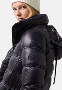 Doudoune bleu marine foncé avec une finition brillante, dotée d'un col montant, d'une capuche, de panneaux texturés et d'un accent de logo rond sur la manche.