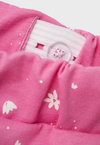 Detail eines rosa Baumwollkleidungsstücks mit einem weißen Blumenmuster, einem elastischen Bund und einem Knopfverschluss mit Schlaufe. Die Textur ist glatt.